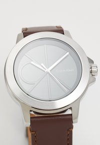 Montre Calvin Klein avec un boîtier en acier inoxydable argenté, un cadran gris, un bracelet en cuir brun et des index heures minimalistes. Elle dispose d'une couronne sur le côté.