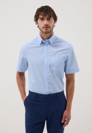 Man met lichtblauw kort mouw overhemd met knopen en donkerblauwe broek, staand met één hand in de zak tegen een effen achtergrond.
