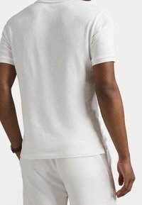 Polo blanc à manches courtes, doté d'une texture douce, de fentes latérales et d'un col simple. Porté avec un pantalon assorti.
