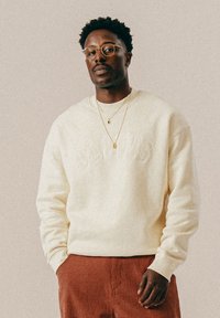 Mężczyzna w okrągłych okularach, złotych naszyjnikach, kremowym swetrze z napisem "BLKVNS" i czerwonych w kratę spodniach, stojący na jednolitym tle.