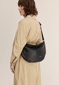 Borsa a spalla in pelle nera con forma arrotondata, caratterizzata da un logo dorato e una tracolla regolabile, indossata sopra un trench coat beige.