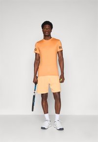 Oranžinė sportinė marškinė su "BG" logotipu, derinami su šviesiai oranžinėmis šortais. Modelis laiko teniso raketę ir žalią teniso kamuoliuką. Balti sportbačiai.