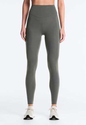 Kvinde iført olivengrønne leggings med høj talje og hvide sneakers, stående med armene afslappet ned langs siderne mod en ensfarvet baggrund.