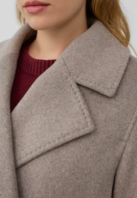 Beżowy płaszcz wełniany z szerokim kołnierzem, z chropowatą powierzchnią i widocznym st stitchingiem, założony na burgundowy sweter dzianinowy.