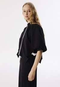 Zwarte cropped sweatshirt met een hoge kraag en een witte rits. Heeft volumineuze mouwen en een gestructureerde stof. Gecombineerd met hoge, zwarte broeken.