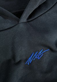 Zwarte hoodie met een zachte textuur en een blauw geborduurd ontwerp op de onderste rechterkant. De stof is glad en iets rekbaar.