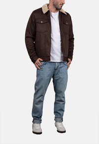 Uomo che indossa una giacca marrone con colletto in shearling beige, una maglietta bianca, jeans azzurri chiari con risvolti e scarpe da ginnastica bianche, in piedi con le mani in tasca.