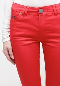 Pantalones de mezclilla rojos ajustados, con un bolsillo frontal adornado con rhinestones y un cierre de botón metálico estándar.