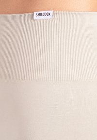 Beige ribgebreide leggings met een gladde textuur en een wit label met het opschrift "SMILODOX" aan de tailleband. De stof lijkt rekbaar en nauwsluitend.