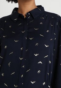 Blusa blu navy con motivi astratti a foglia dorata, con bottoni sul davanti e colletto classico indossata da una persona.