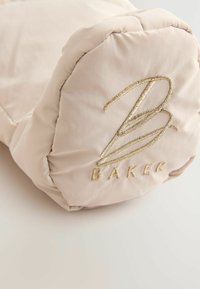 Sac en tissu beige rembourré avec une finition brillante, présentant une broderie dorée du "B" et du texte "BAKER", des bords arrondis et une texture douce.
