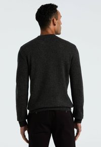 Von hinten sichtbarer Mann, der einen dunkelgrauen Strickpullover und schwarze Hosen trägt, vor einem schlichten hellen Hintergrund.