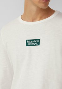 White long-sleeve t-shirt made of soft fabric, featuring a green rectangular label with white text and a floral icon at the chest.