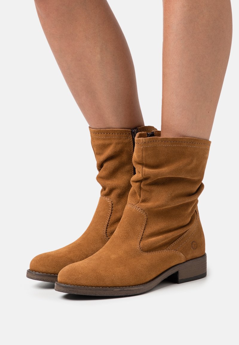 Bottes marron en daim mi-mollet avec fermeture éclair latérale, portées sur des jambes nues, dotées de bas talons empilés et d'un design légèrement affaissé.
