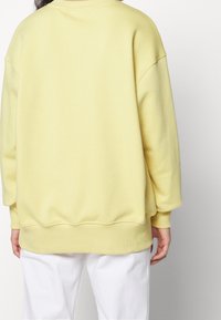 Žlutá mikina s volným střihem, žebrovanými manžetami a lemem. Měkká textura, široké rukávy a kulatý výstřih. Nnošená přes bílé kalhoty.