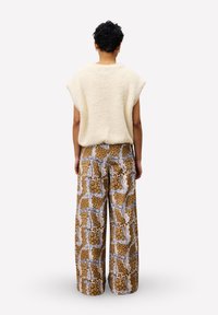 Personne avec des cheveux courts et bouclés, portant un haut crémeux sans manches en peluche et un pantalon large à motifs marron, se tenant de dos.