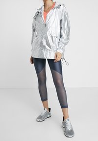 Chaqueta ligera plateada con capucha, combinada con leggins marinos que presentan paneles transparentes y zapatillas deportivas grises. Diseño limpio, estilo funcional.