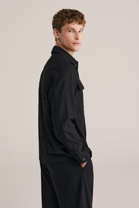 Camicia nera, modello slim fit, realizzata in tessuto liscio. Presenta una tasca sul petto e polsini con bottone, mostrata su un modello di lato.