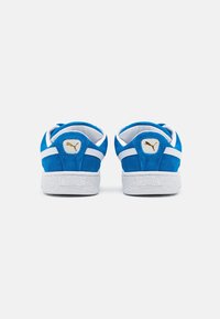 Zapatillas de ante azul con acentos blancos, parte trasera redondeada y suela de goma blanca texturizada. Presenta un logo dorado en el talón.