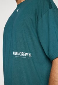 Zöld pamut póló kerek nyakkal. A bal oldalon található fehér felirat: "PUMA CREW '48". Puha tapintású és laza szabású.
