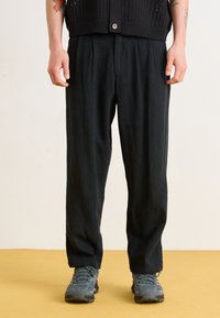 Black, loose-fit trousers with pleated front, made from lightweight fabric, paired with gray hiking shoes. Features a relaxed, flowing design.