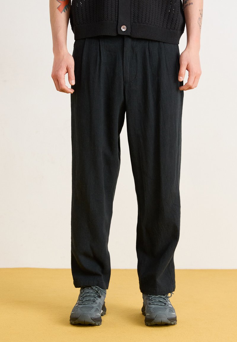 Black, loose-fit trousers with pleated front, made from lightweight fabric, paired with gray hiking shoes. Features a relaxed, flowing design.