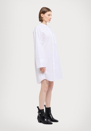 Bílá oversized košilová šaty s vysokým límcem, dlouhými rukávy a předním knoflíkovým zapínáním; nošeno s černými kotníkovými botami se špičatou špičkou.