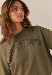 Olivgrünes Baumwoll-T-Shirt mit einem auffälligen "COPENHAGEN"-Aufdruck in Schwarz auf der Brust. Klassischer Rundhalsausschnitt und lässiger Schnitt.