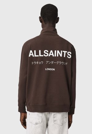 Persoon die een donkerbruine sweatshirt draagt met "ALLSAINTS", Japanse tekst en "LONDON" in het wit gedrukt op de achterkant, gecombineerd met lichte jeans.