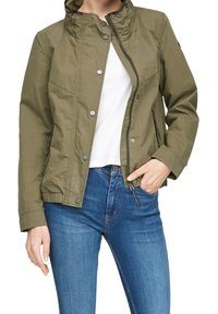 Osoba w oliwkowo-zielonej kurtce na zamek, białej koszuli i niebieskich dżinsach, z jedną ręką w kieszeni na białym tle.