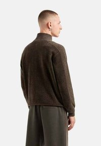 Maglione tricot marrone con un colletto alto, tessuto strutturato e vestibilità rilassata. Presenta polsini e orlo a coste, indossato sopra pantaloni grigi chiari.