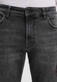 Primo piano della sezione centrale che indossa jeans neri sbiaditi con chiusura a bottoni e zip, tasche anteriori e una camicia bianca infilata dentro.