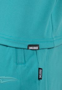 Teal Sportbekleidung mit glatter Textur, versehen mit einem Logo am Bund und einem Etikett mit "SMILODOX" in Schwarz an der Seite.