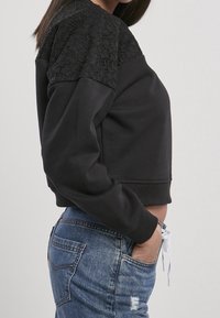 Femme en haut noir à manches longues avec épaules en dentelle et jean bleu déchiré, debout de côté avec une main dans la poche.