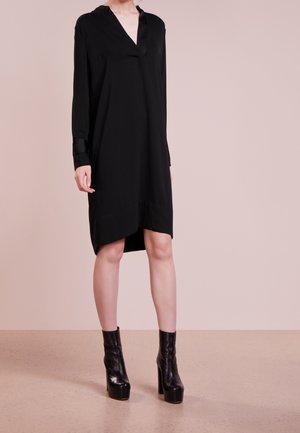 Kvinde iført en sort V-hals kjole med lange ærmer kombineret med sorte læderankelstøvler med høje hæle, stående på en beige gulv.