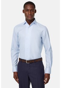 Lichtblauwe overhemden met een subtiele geblokte print, lange mouwen en een klassieke kraag. Draagt een bruine suède riem en marineblauwe broek.