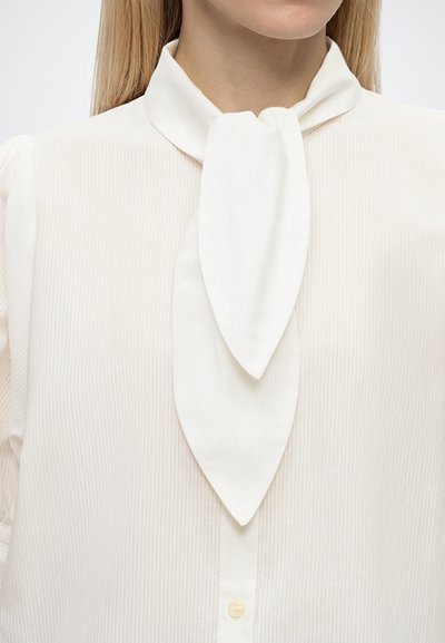 Femme portant une blouse blanche avec un col haut et un détail de foulard noué au cou, montrant le haut de la poitrine et le bas du visage.