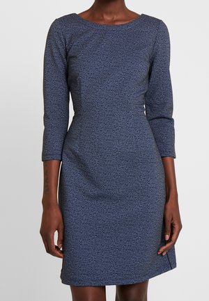 Kvinde iført en tætsiddende, blå, tekstureret kjole med trekvartlange ærmer og bådudskæring, stående mod en ensfarvet baggrund.