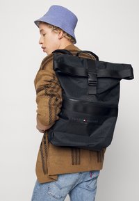 Sac à dos noir à rabat en matériau synthétique texturé, avec une poche zippée à l'avant et un détail de logo. Porté sur un pull à motif marron.