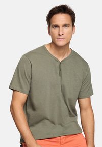 Hombre con cabello corto y castaño, vestido con una camiseta henley de manga corta color verde oliva y pantalones color coral, mirando al frente con expresión neutra.