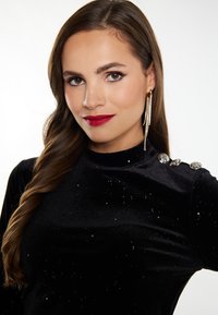 Top in velluto nero con scollatura alta, decorato con accenti argentei sulla spalla e una leggera lucentezza. Capelli lunghi acconciati in onde morbide.