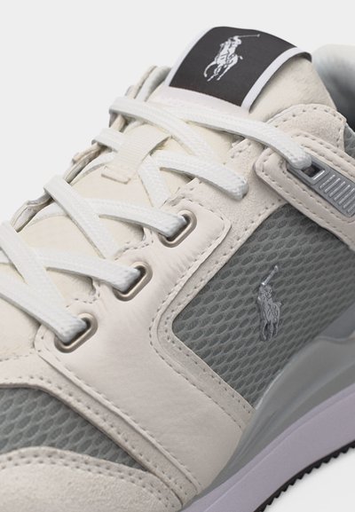 Primer plano de una zapatilla beige y gris con cordones blancos, panel lateral de malla y el logo de Polo Ralph Lauren en la lengüeta y el lateral.