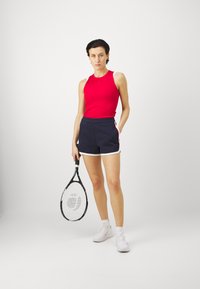 Rød ermeløs tanktop, marineblå shorts med hvite kanter, holder en svart tennisracket, iført hvite joggesko. Enkelt hvitt bakgrunn.