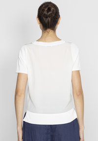 Biała bluzka z krótkim rękawem o gładkiej teksturze, z przezroczystą tylną częścią i solidnym białym dołem. Podkreślona subtelnymi detalami wzdłuż szwów.