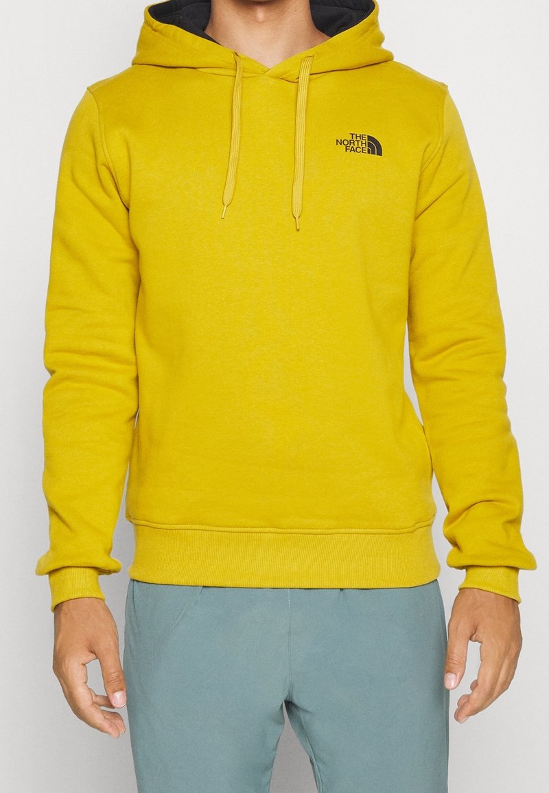 Homme portant un sweat à capuche The North Face jaune vif avec un logo noir sur la poitrine et un pantalon bleu clair, debout devant un fond uni.