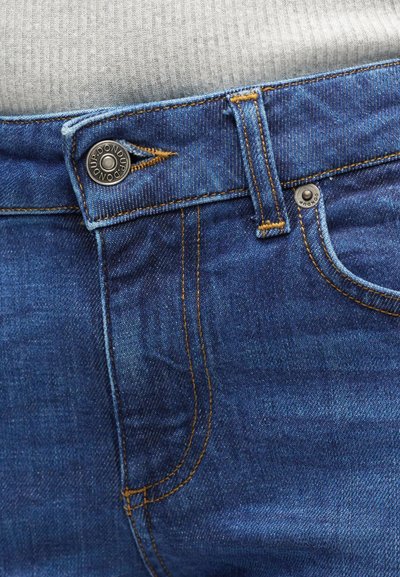 Gros plan sur la ceinture d'un jean en denim bleu avec un bouton métallique et des coutures visibles, porté avec un haut côtelé gris clair.
