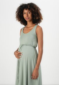 Mulher grávida com cabelo encaracolado, vestindo um vestido sem mangas de cor verde claro, segurando suavemente a barriga, de pé contra um fundo branco.