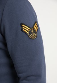 Granatowa bluza z suwakiem z naszywką z złotymi skrzydłami, kotwicą i napisem "DreMarin". Materiał ma miękką, teksturowaną powierzchnię.