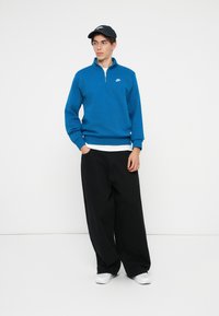 Μπλε φούτερ με φερ�μουάρ που είναι φτιαγμένο από βαμβάκι, με λευκό λογότυπο στο στήθος, συνδυασμένο με χαλαρούς μαύρους παντελόνες και λευκά αθλητικά παπούτσια.