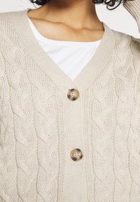 Žena v krémovém kardiganu s pleteným vzorem a dvěma velkými hnědými knoflíky, pod nímž má na sobě bílý top, který odhaluje límec a horní část hrudníku.